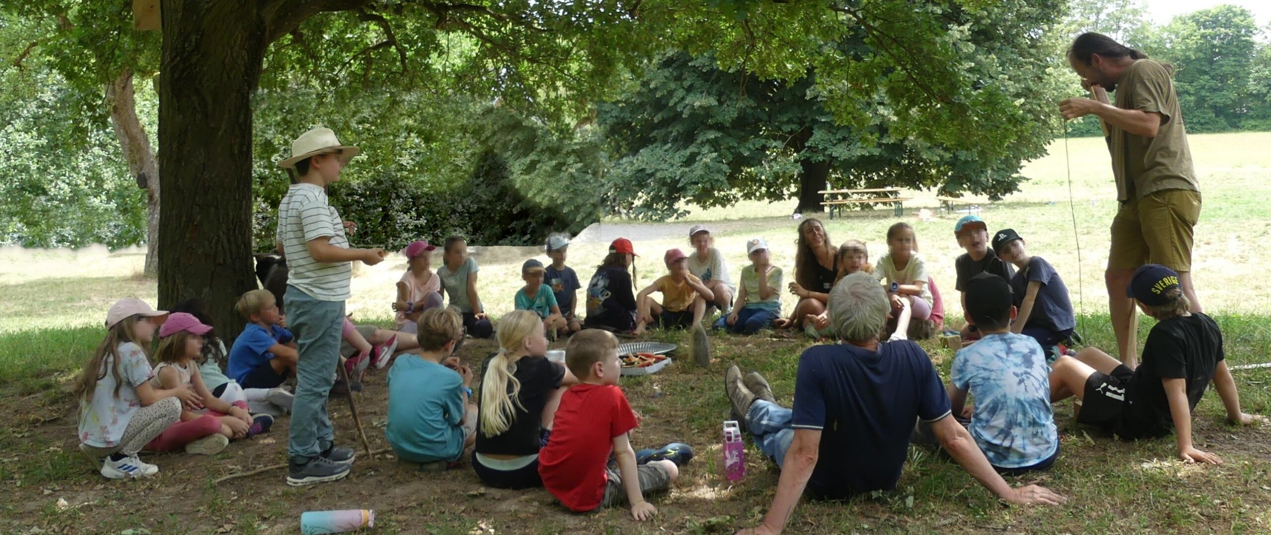 Kinder und Erwachsene sitzen beim Steinzeit-Camp im Kreis im Schatten einer großen Eiche, ein Junge mit Hut steht und erzählt